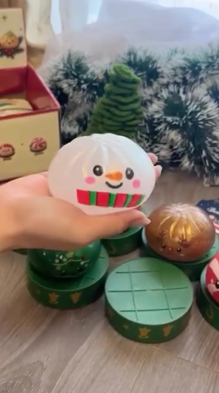 A hand holds a translucent white snowman stress ball featuring a cute smiling face, orange nose, and a red and green striped scarf design against a holiday background.