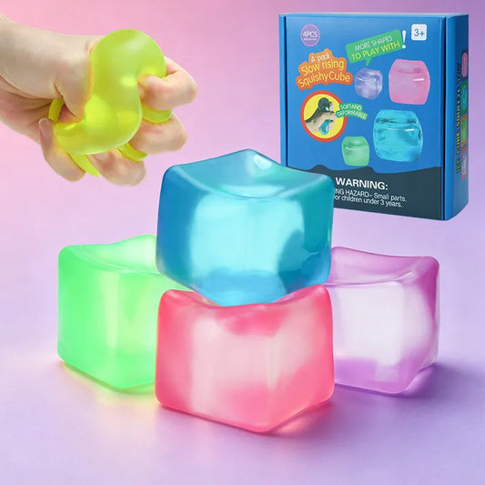Four colorful translucent slow-rising squishy cubes used as fidget toys for stress relief. A hand demonstrates the soft texture of a lime green cube, accompanied by blue, pink, and purple versions. Retail packaging sits in the background.