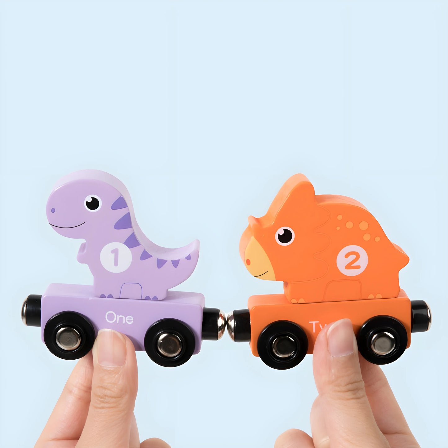 Two magnetic dinosaur-shaped toy cars, one purple with the number 1 and one orange with the number 2, held by a person against a light blue background.