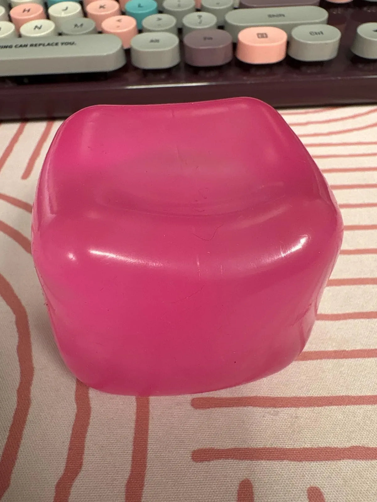 Glossy bright pink silicone charging dock stand with rounded edges sitting on a striped mat. A mechanical keyboard with pastel keycaps is visible in the background. Modern desk accessory for organizing electronics.