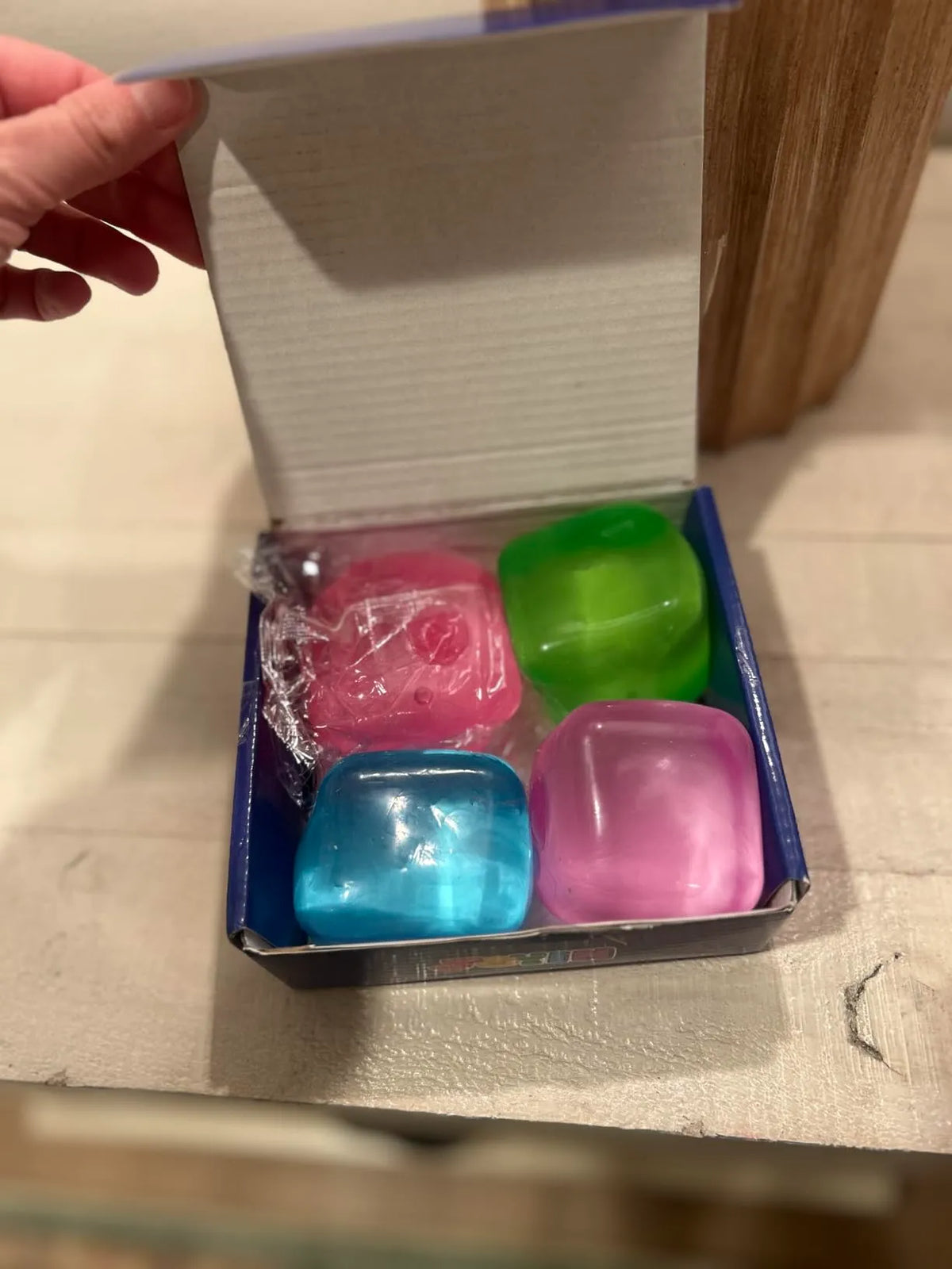 Blue box opened by hand reveals four glossy, translucent cubes in pink, green, blue, and purple. One pink cube features a molded character face. Colorful items sit in the package on a wooden surface.