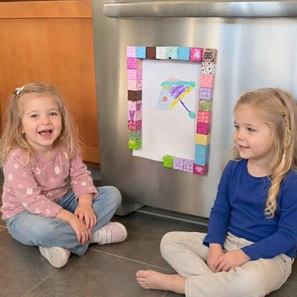 Two children sitting on the floor in front of a refrigerator with a colorful magnetic frame.